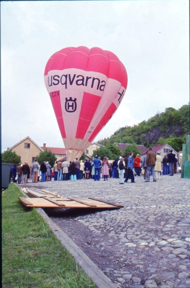 Den rödvita ballongen "Örnen II Gränna" med reklam för "Huskvarna" står fylld på Gränna torg. En folksamling runt ballongkorgen.