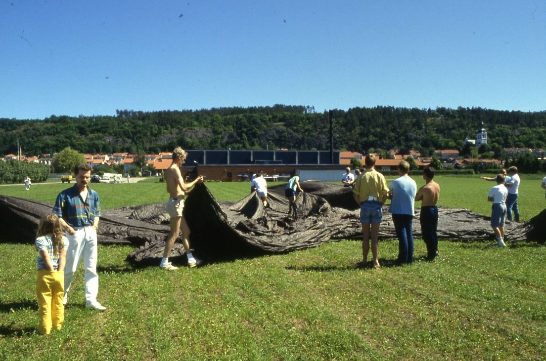 Ett stort brunt ballonghölje ligger utlagt på en gräsplan. Det är kopian av Andrées ballong Örnen från Jan Troells biofilm "Ingenjör Andrées luftfärd". Barn och vuxna runtomkring.