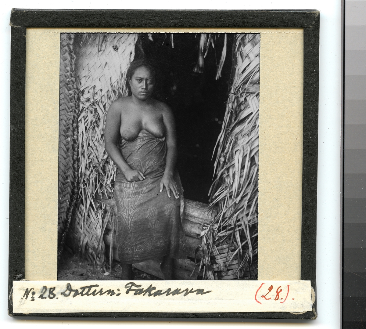 Bilden visar en kvinna från ursprungsbefolkningen på Fakarava klädd i en traditionell dräkt som står framför en hydda. Enlig Etnografiska museets bildinformation är det dottern Taroi.