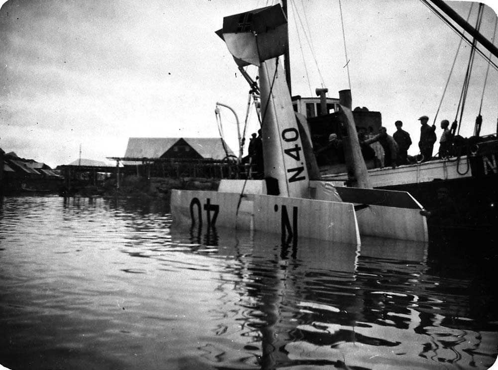 Ett sjøfly som blir heist opp av vannet etter et havari, Saaski "Måsen". En båt med flere personer ombord. Bygninger i bakgrunnen.