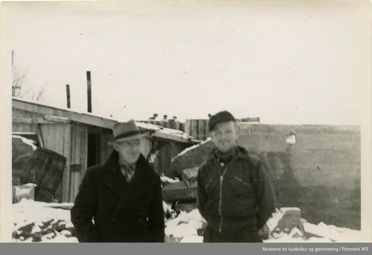 To menn står foran et provisorisk bygg, et gjerdet og en grunnmur. Bildet er trolig tatt på Hauen i Hammerfest. Mennene smiler. Det er snø på bakken.