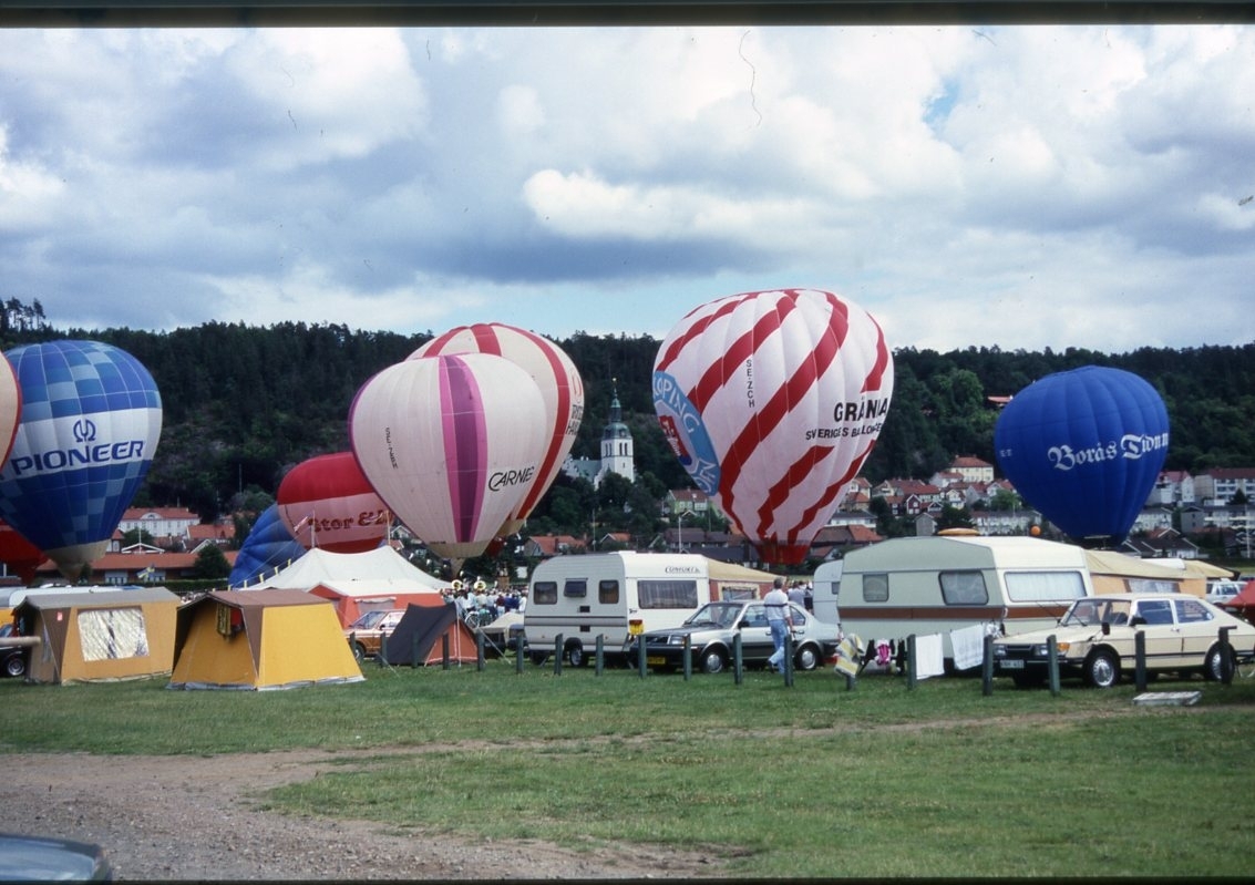 Luftballonger fylls och lyfter från en gräsplan. En stor publik. Husvagnar och tält i förgrunden.