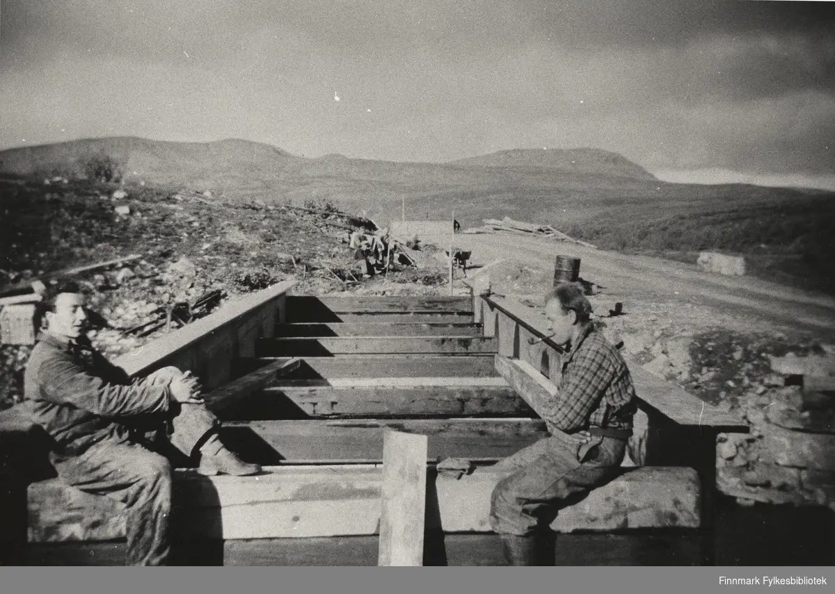 Krigskadeutbedringer i Finnmark 1946. Portrettfotografi av to menn som sitter på en påbegynt bru på Ifjordfjellet.