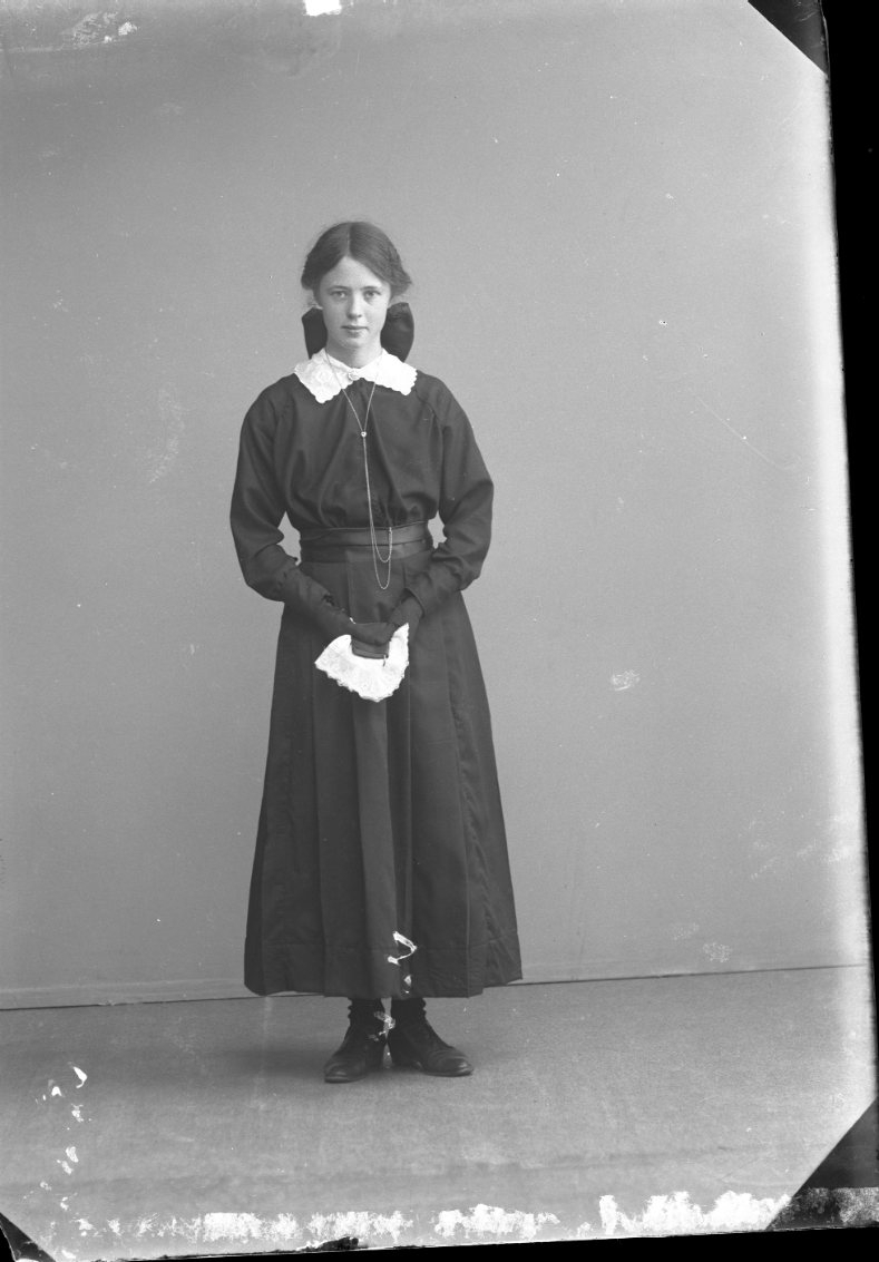 Porträtt av en flicka i mörk klänning. Hon har svarta handskar och håller i en ljus näsduk och en liten bok, möjligen en psalmbok. Konfirmationsbild?