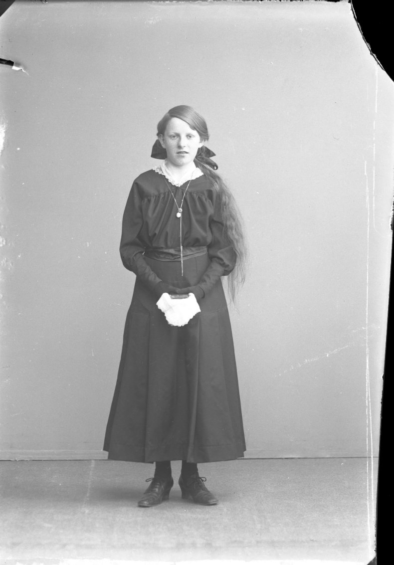 Porträtt av en flicka i mörk klänning. Hon har svarta handskar och håller i en ljus näsduk och en liten bok, möjligen en psalmbok. Konfirmationsbild?