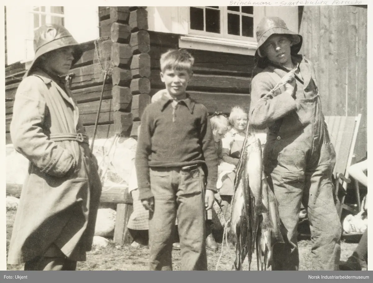 Barn foran laftet hytte på Briskeroe, Møsstrond. To gutter iført sydvest viser dagens fiskefangst. Mellom de to guttene. Fra venstre: Esben Poulsson, Anton Martin Schweigaard og Magnus Poulsson