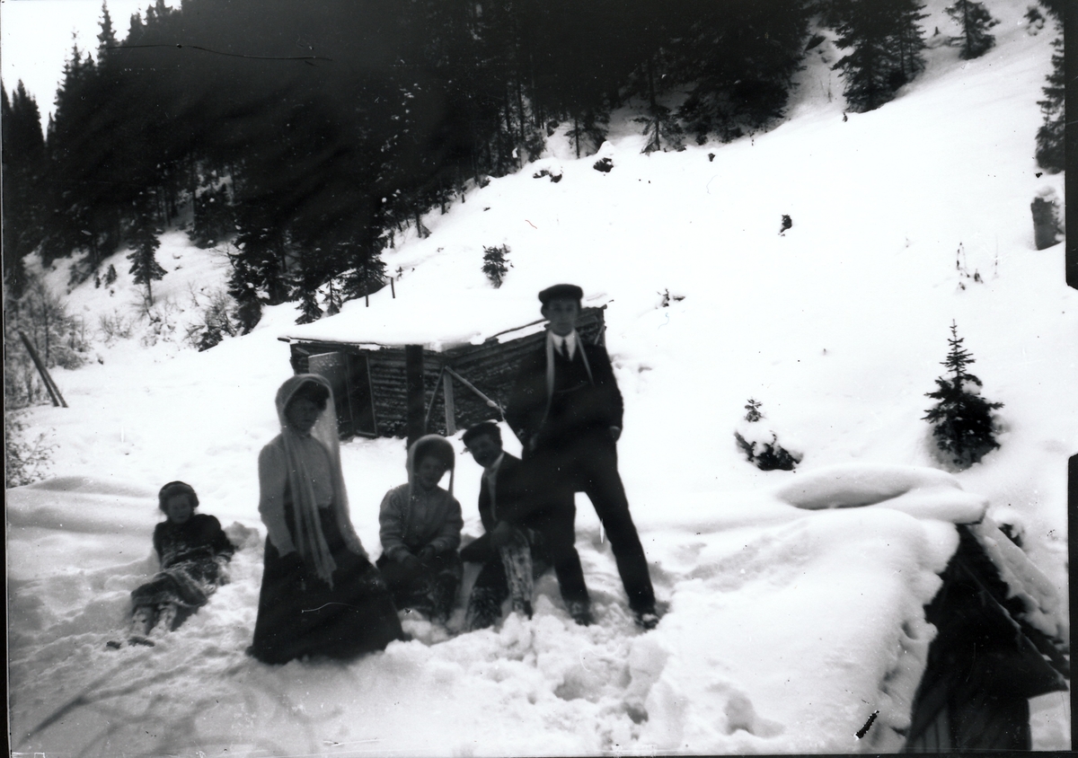Medlemmer og venner av Rydgrenfamilien på et snødekket tak. Harald Rydgren er mannen som sitter i snøen og Signe Rydgren er kvinnen som sitter ved siden av ham. I bakgrunnen et uthus med snødekte tak. De to kvinnene i bildet har lange skjerf over luer.
