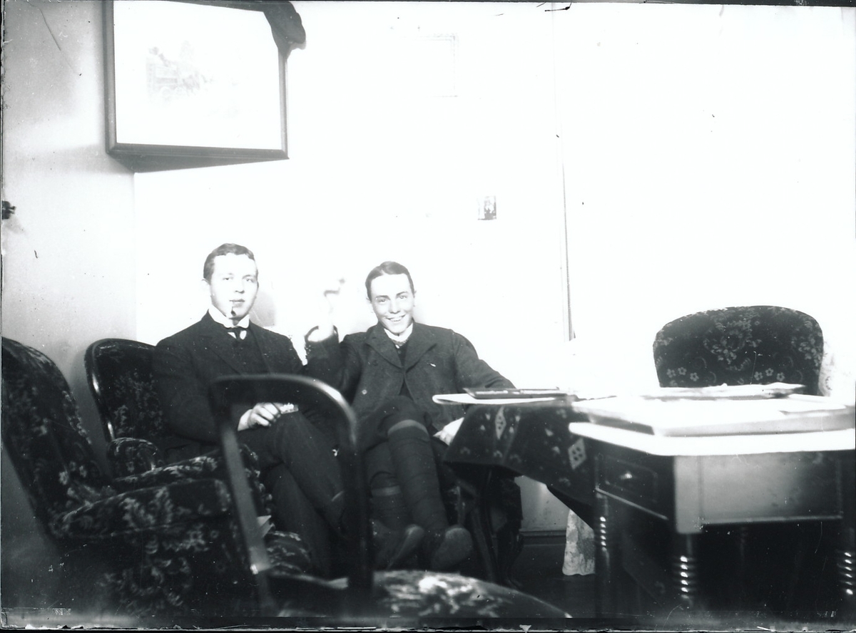 To unge menn sitter i dype stoler, Harald Rydgren til venstre og han røyker pipe.