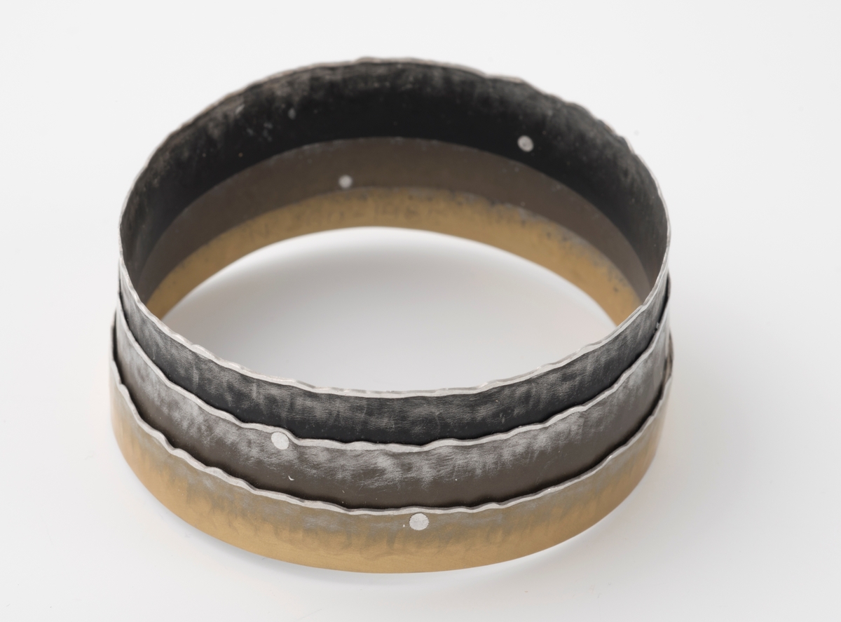 Armbånd bestående av tre flathamrede ringer som er lagt utenpå hverandre og festet sammen med små, blanke nagler. Ringenes ene side er svakt bølgeformet og aluminiumsfarget. Ringenes farger er mørk sotfarget, brun og lys gylden.