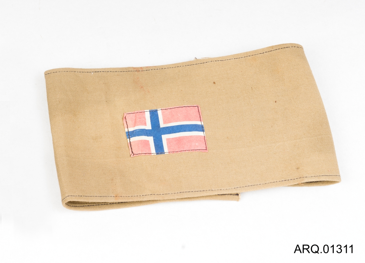 Armbind med påsydd norsk flagg. Ved nærmere ettersyn er det papirflagg som er sydd og dette armbindet er trolig laget som utstillings-gjenstand (rekvisita).