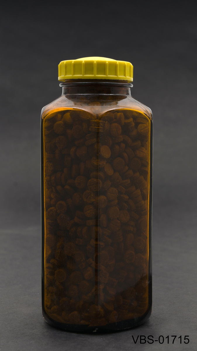 Sekskantet laboratorieglassflaske i ravbrun farge. Nederst på flasken er det en lettelse med påskriften 500.
Inneholder brune tabletter.
Gul bakelitt skrulokk.
