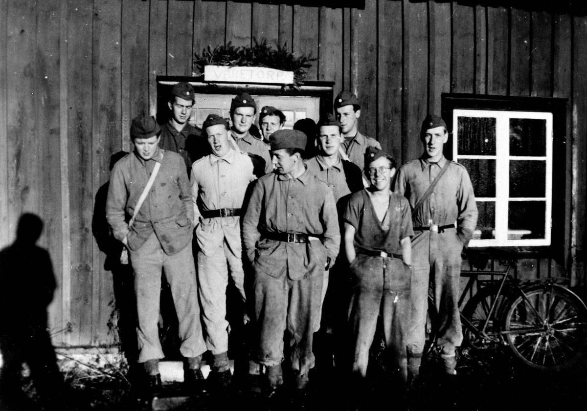 Gruppbild av 10 män, i uniform, som står framfår en ingång till ett trähus. Ovanför dem en skylt med text "Varetorp" eller "Väretorp".
Beredskapen/rekryten. Skördepermission.