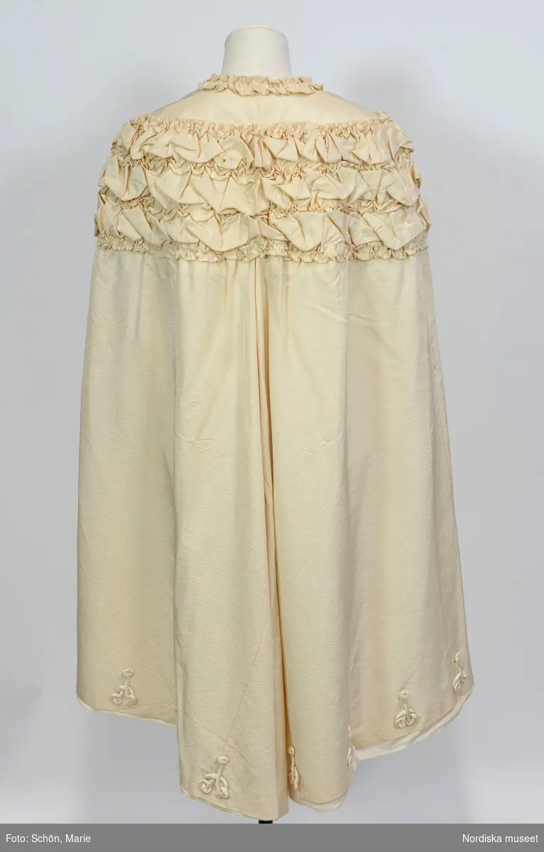 Ordered by Ingrid Bergström née Anjou (1856–1948), married to cabinet minister David Bergström, a major supporter of the right to vote for all. Delivered 1906. White silk cape with decorative agraffs and tassels. Worn by Ingrid Bergström at the christening of 
Prince Gustaf Adolf in the Royal Chapel in 1906.