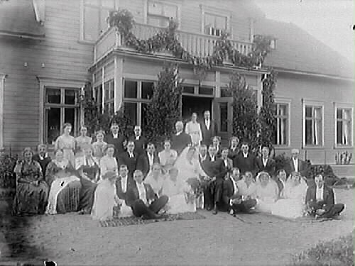 Brudpar med bröllopsgäster samlade utomhus vid lövprydd veranda till ett stort trähus. Balkongen ovanpå pryds av en girland. Personerna längst fram sitter på mattor i sanden.