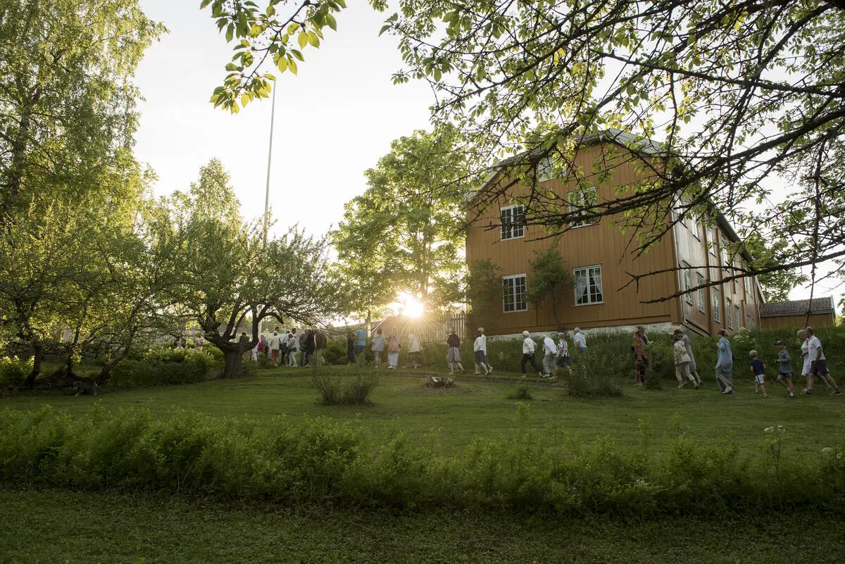 Tverrenden av et det store okergule hovedhuset på amtmannsgården Steinberg på Toten. Lav sol i mot, og en lang rekke av mennesker på vei gjennom den grønne parken foran huset.