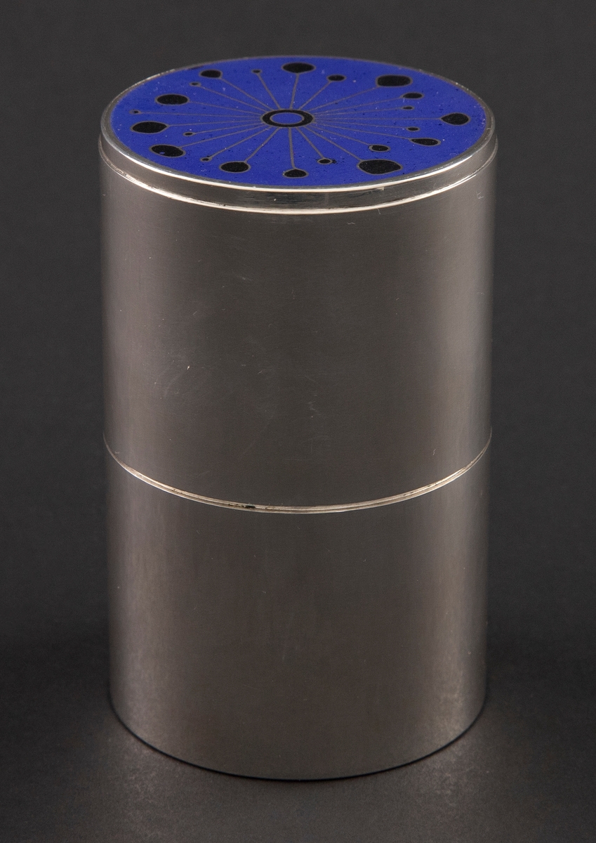 Sylinderformet sigrettboks i sølv, bestående av to deler. Den øverste delen fungerer som et overfalset lokk, og er dekorert med opak blå- og sortfarget cellemalje. Dekoren viser en geometrisk figur med radiære stråler ut fra et sirkelformet midtpunkt. Resten av boksen er uten dekor.