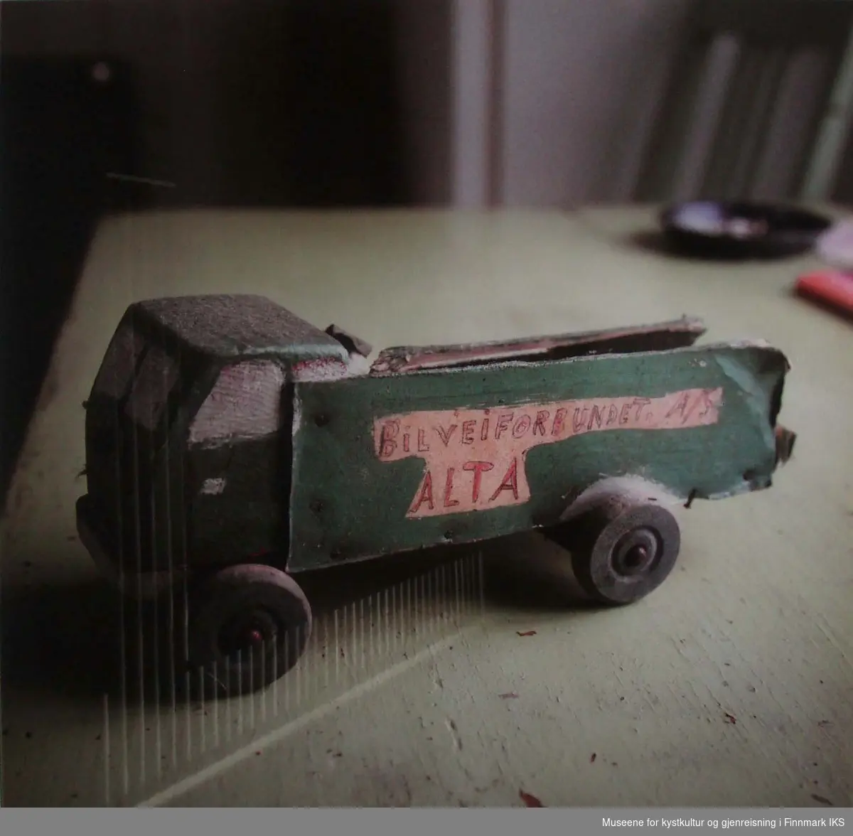 Et hjemmelaget leketøy står igjen i et fraflyttet gjenreisningshus. Leketøyet er en lastebil av grønn malt tre. Sideveggen er av papp med påskriftet "Bilveiforbundet. A/S Alta".