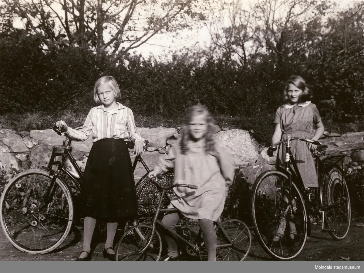Ulla till höger på sin cykel vid muren. I mitten Bibbi samt till vänster en kusin.

Fotografi taget i september 1931 vid sommarbostaden Strandlyckan vid Landvettersjön, Slamby 1:4 i Landvetter socken. Huset övertogs senare av Stiftelsen Enandergården, Härlanda församling, på 1950-talet.