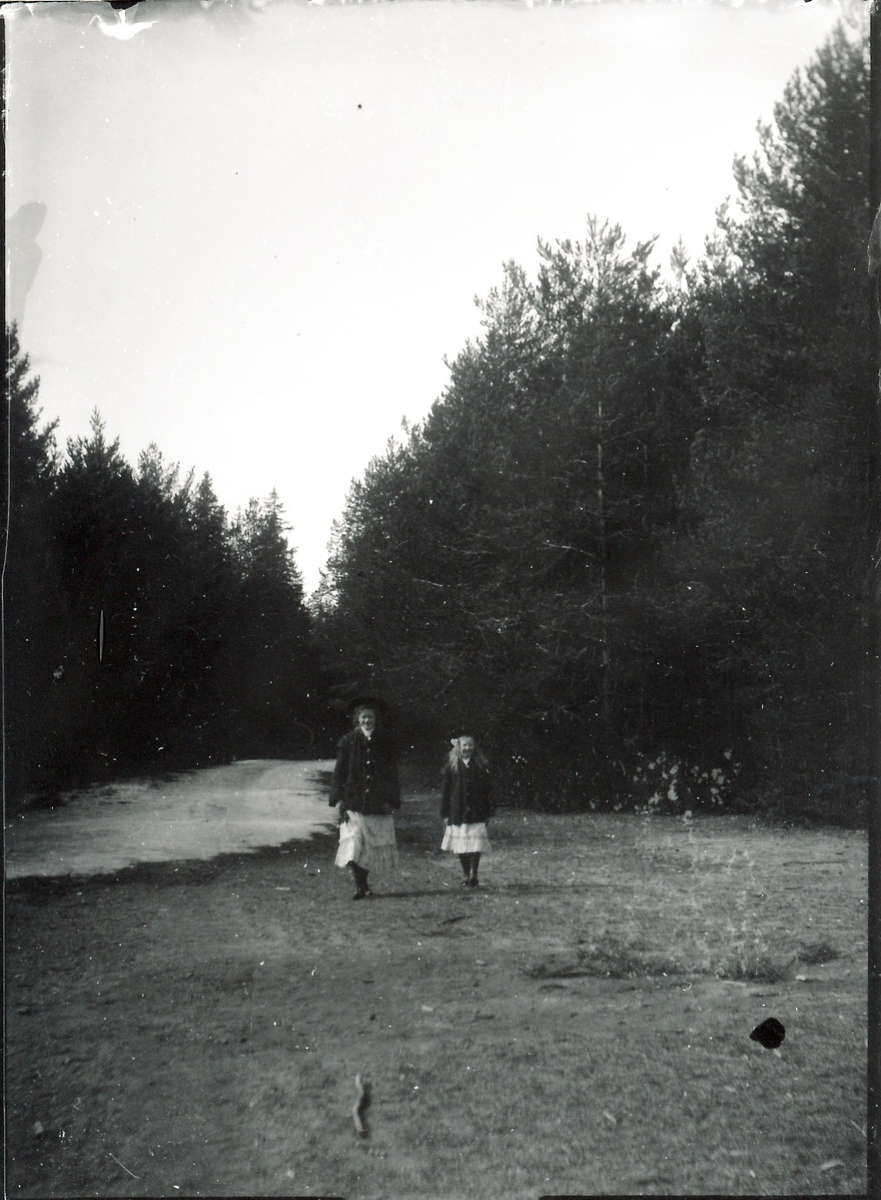 Søstrene Signe og Eivor Rydgren på tur. Jentene går på en vei i skogsterreng, likt kledd med hatter, vide jakker og volangskjørt. Snøflekk til venstre.