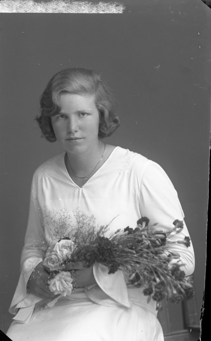 Porträtt av en ung kvinna i ljus klänning och blomsterbukett i famnen. En konfirmand?