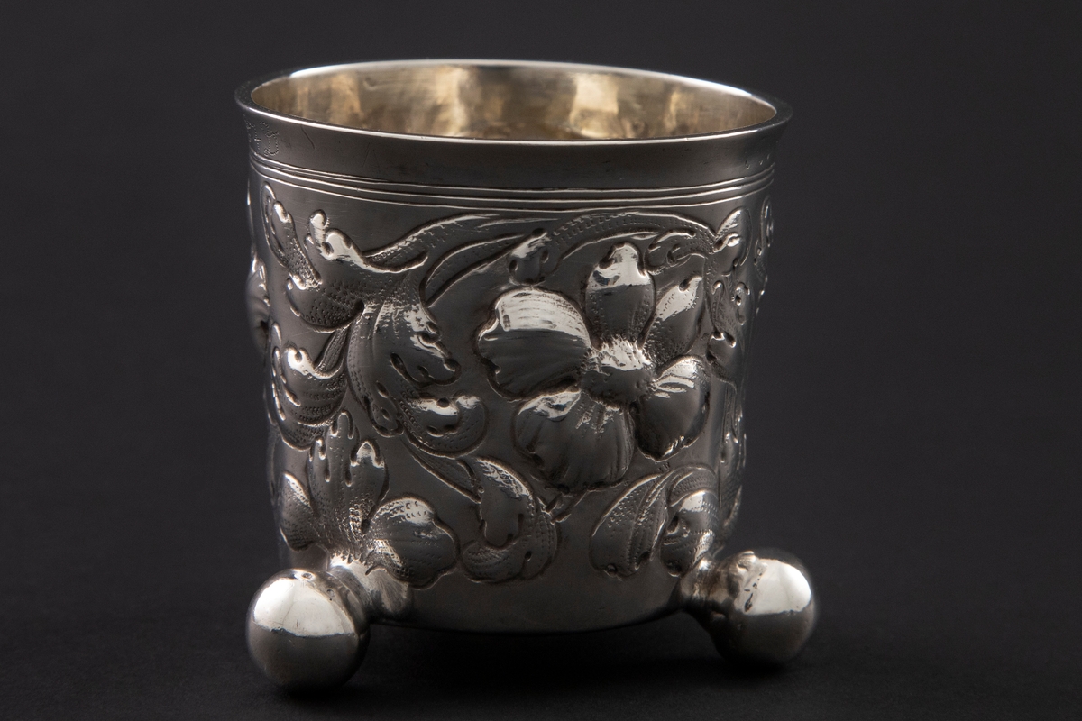 Sølvbeger med svakt konisk form som hviler på tre kuleben. Korpus er dekorert med drevet, gravert og siselert blomsterornamentikk
