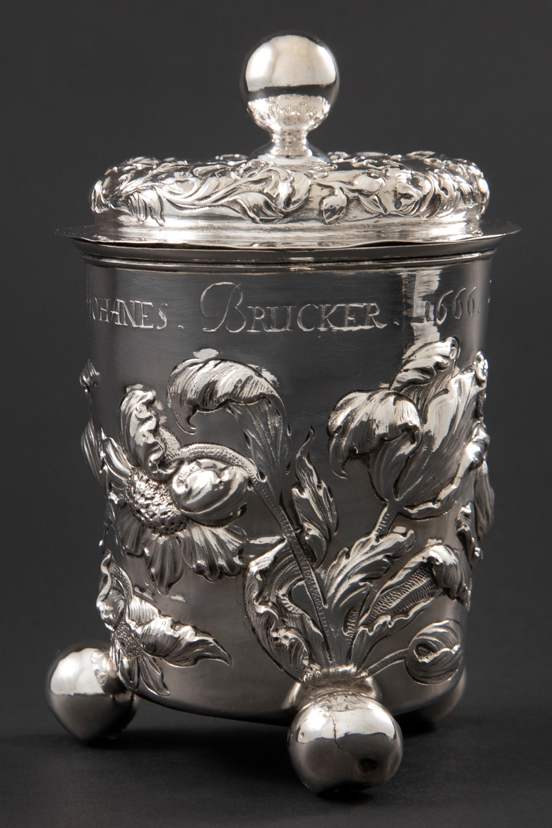 Lokkbeger i sølv med svakt konisk form som hviler på tre kuleføtter. Lokknappen er utformet som en kule. Korpus og lokk er dekorert med rikt drevet blomsterornamentikk.