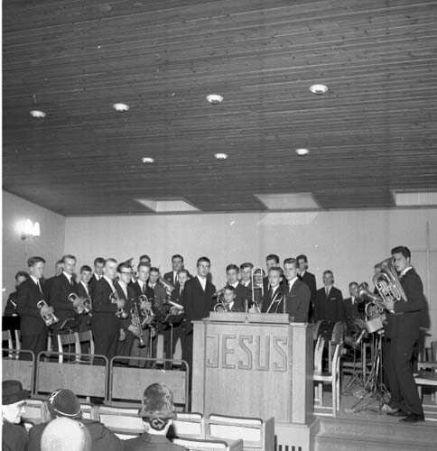 Scen med ett talarbord sett framifrån. Det står Jesus på talarbordet. En blåsorkester står på scenen snett mot publiken. De har sina instrument i händerna.