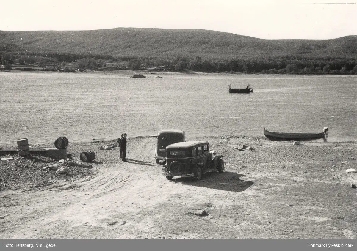 Seida fergested. Seida ferge er på tur over Tanaelva, 1948. Til venstre ser man et fundament for bukkebrua som ble satt opp i 1939. Ved elva ligger en elvebåt. På land står to biler. To menn står ved siden av bilene.