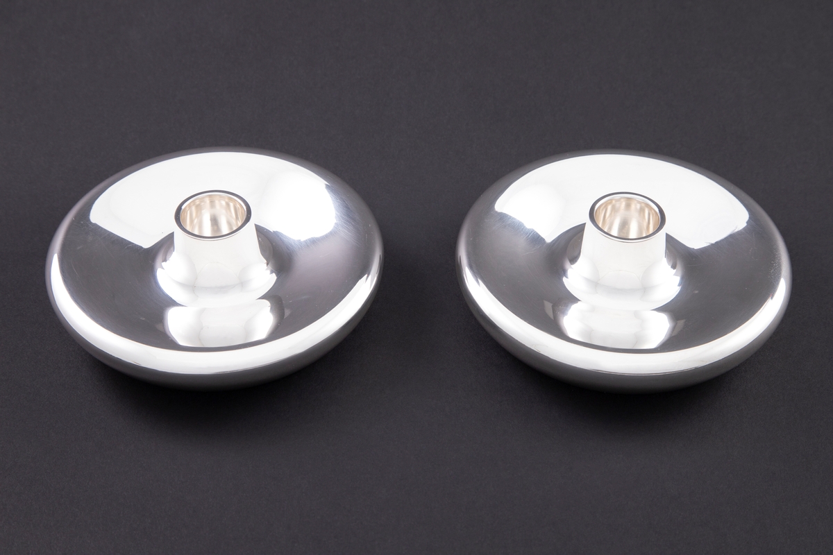 To lysestaker i sølv. Lav, sirkulært korpus med konvekst midtparti og sylindrisk lyspipe.