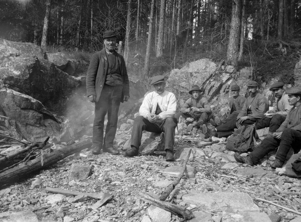 Fløterbasene Per Fossum (1846-1925) og Edvard Lapstun (1875-1950), fotografert sammen med fem andre fløtere i strandsona på Tjuvholmen i Glomma, ved Braskereidfoss i Våler i Hedmark. Fossum sto, Lapstun satt på en steinblokk. Til høyre for ham satt en del andre fløtere.