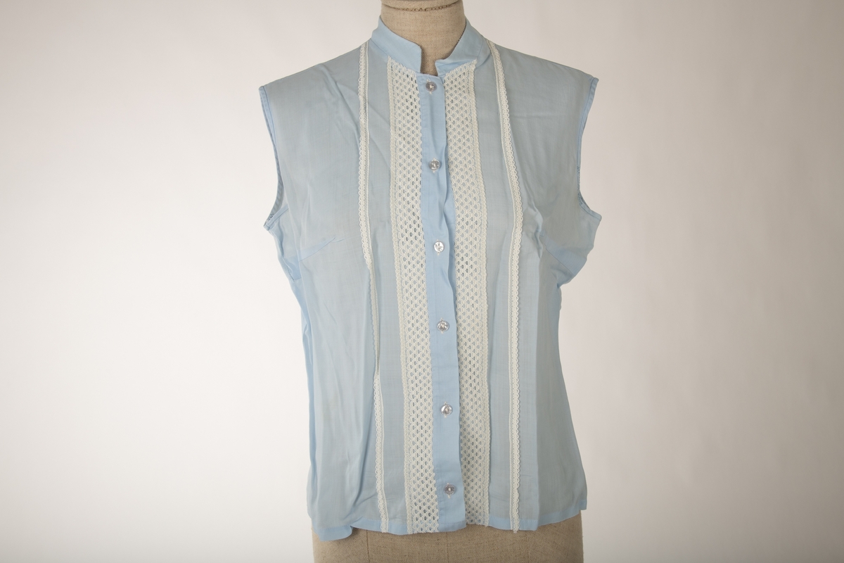 En lyseblå ermeløs bluse, med hvite kniplede bånd sydd på i front. Krage uten brett og knapper og knappehull foran.