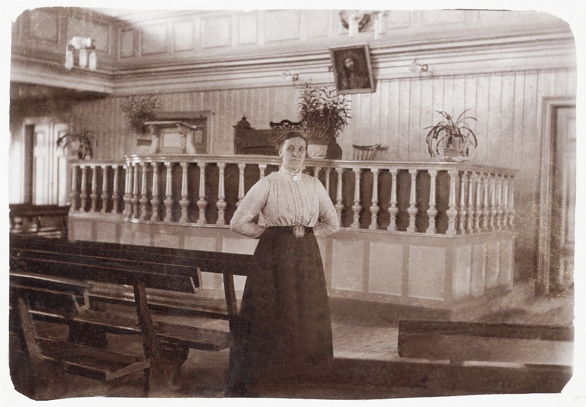 En medelålders kvinna står vid en bänk i Växjö missionskapell, ca 1905.