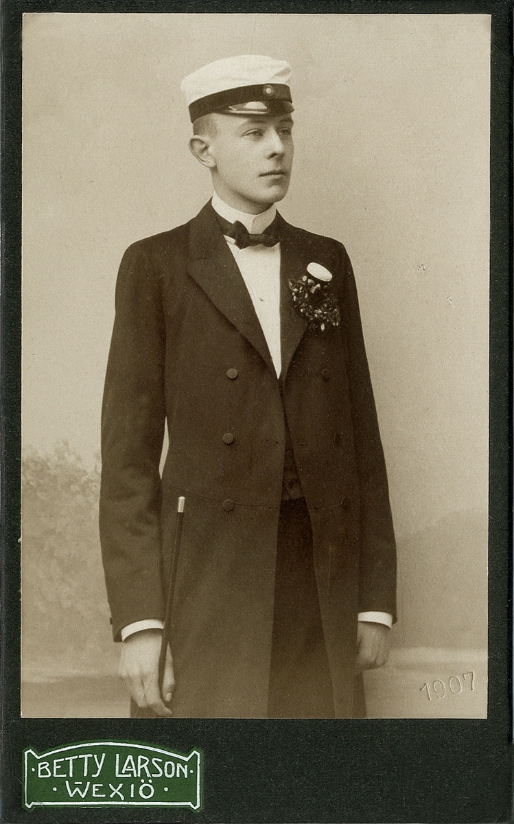 En ung man i redingot med väst, stärkkrage och studentmössa. 
Knäbild, halvprofil. Ateljéfoto 1907.