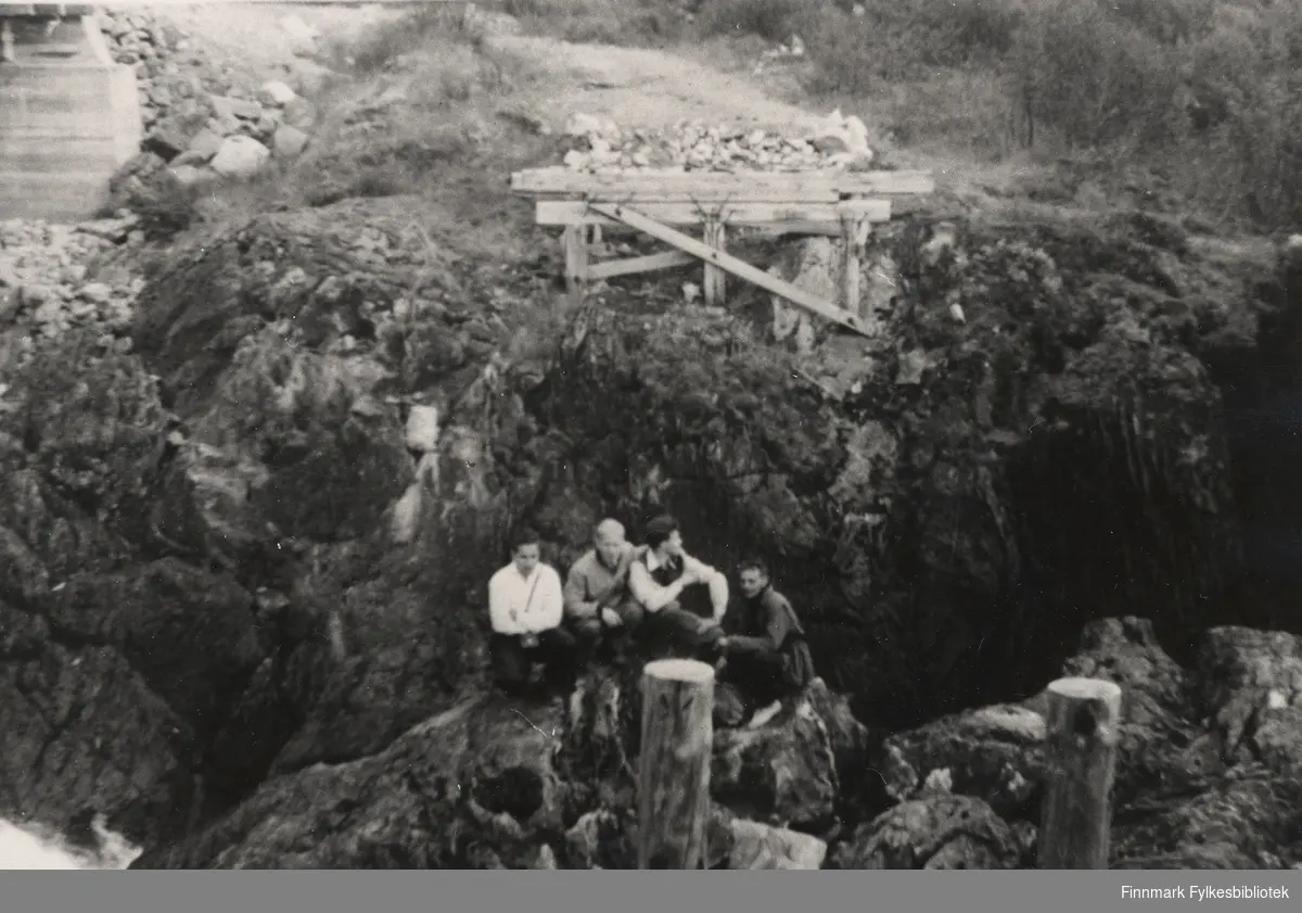 Rester etter den provisoriske bruen over Adamselva ved Adamsfossen. Fire menn sitter tett ved elvekanten.