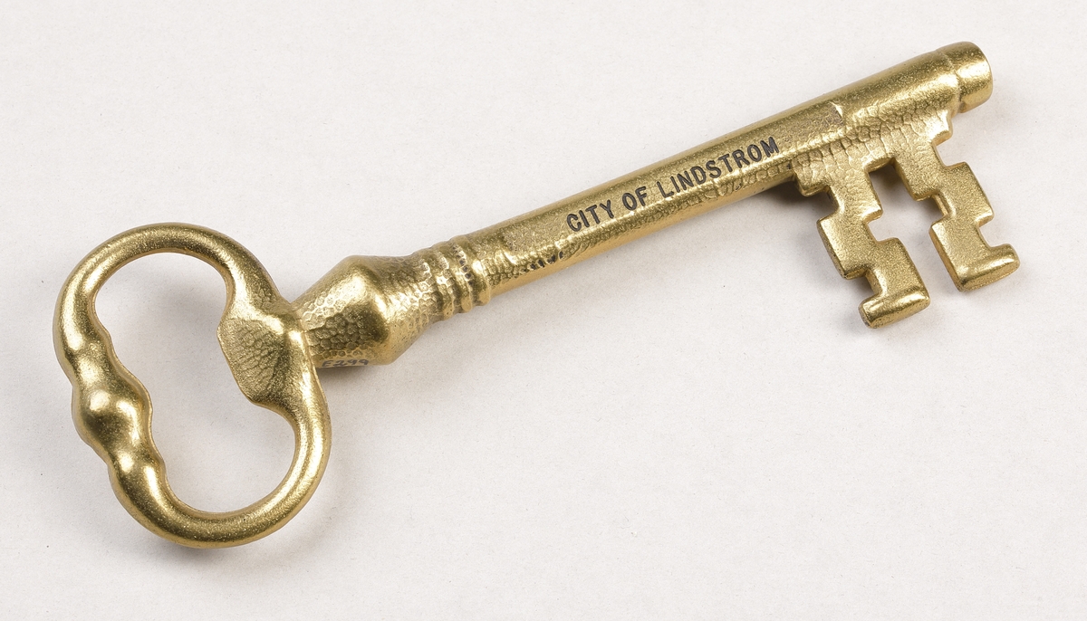 Nyckel i gjuten mässing med ax liknande ett kors. På ena sidan av nyckeln finns texten "VALKOMMEN" och på andra texten "CITY OF LINDSTROM". De präglade texterna är fyllda med svart färg.