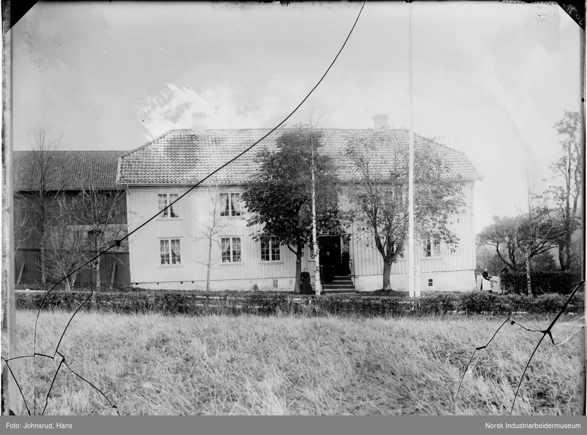 To etasjers bolig med en gruppe mennesker i inngangspartiet, foran huset er det tre trær og en flaggstang. I forgrunnen er det et jorde og bak boligen er det en bygning.
