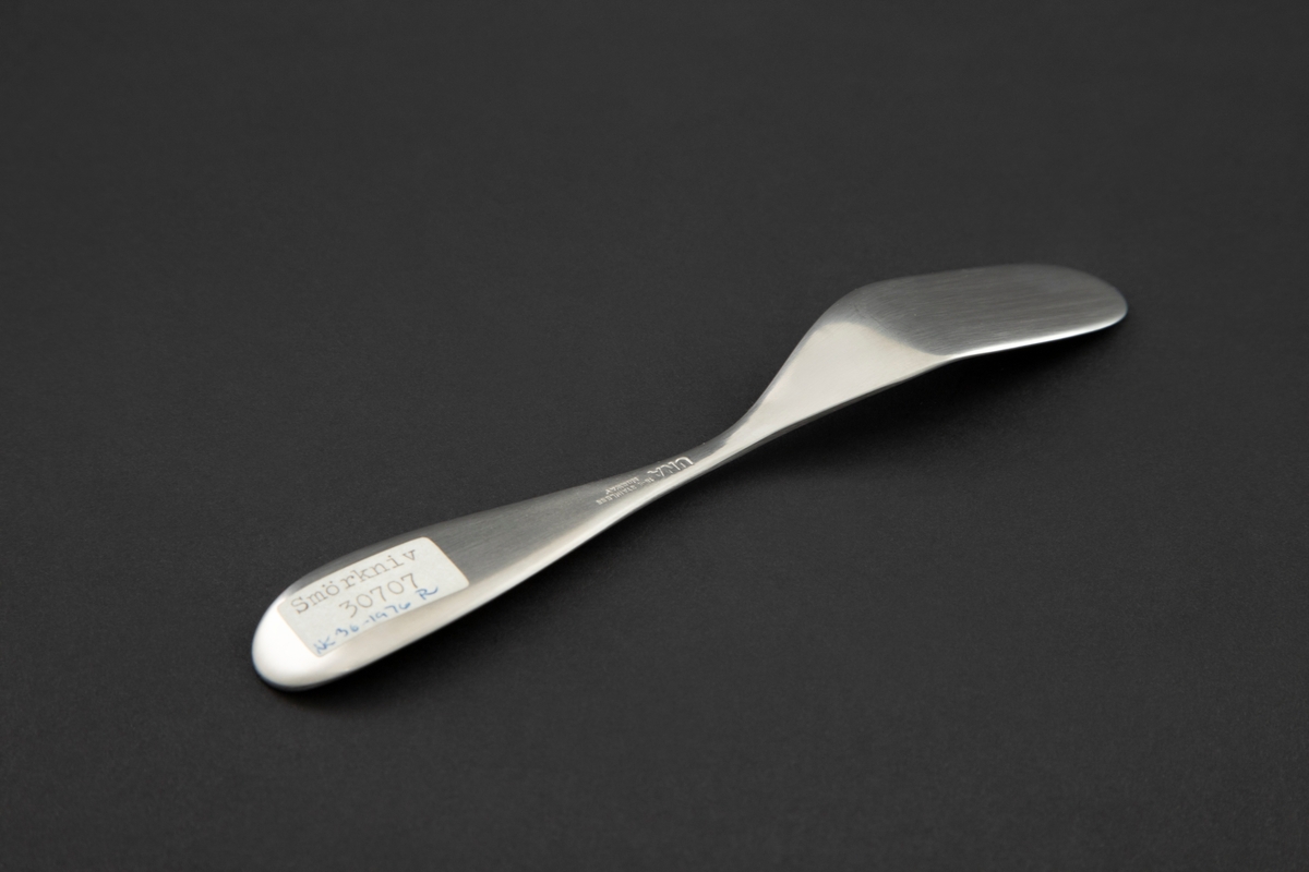Smørkniv i mattpolert rustfritt stål. Ovalt knivblad med rette kanter som skråner oppover. Smal legg og dråpeformet skaft med oppbøyd, avrundet ende.