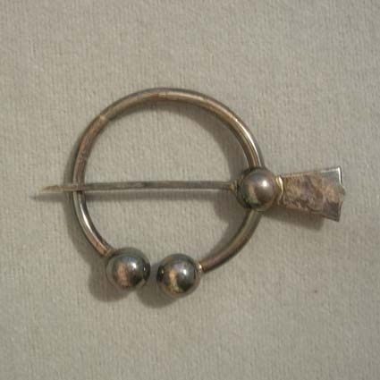 Sölja i silver att fästa ihop Västerbottensdräktens halskläde med. Öppen ring, avslutas med en kula i vardera änden. Fästad i ringen sitter 55 mm lång nål som kan löpa fritt över ringen.