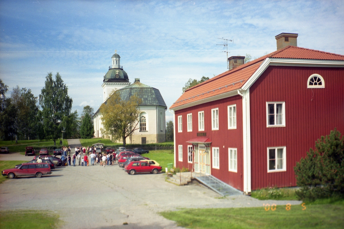 Vy över parkeringen vid Färila kyrka och församlingsgård, 5 augusti 2000. Där står en folksamling vid bilarna.