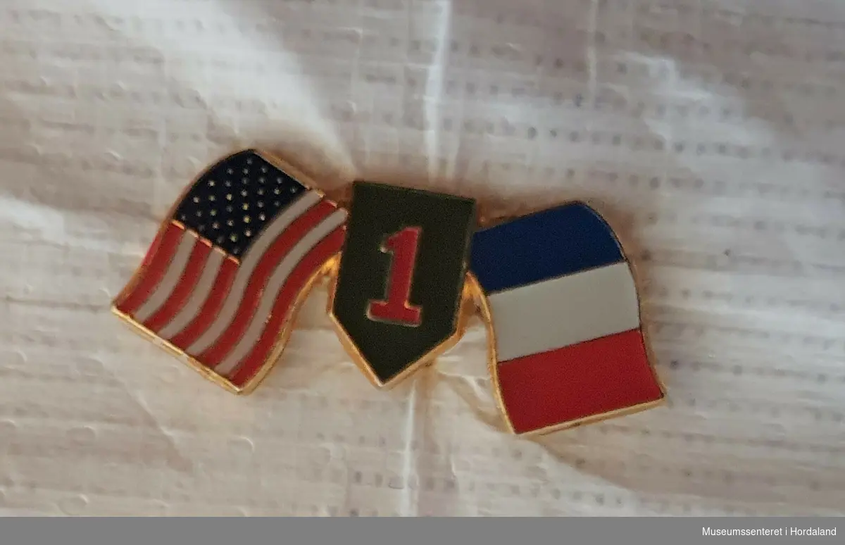 Jakkemerke, pin med to flagg, USA og Frankrike, som vaier på hver side av et merke, rødt ettall på grønn bunn. 