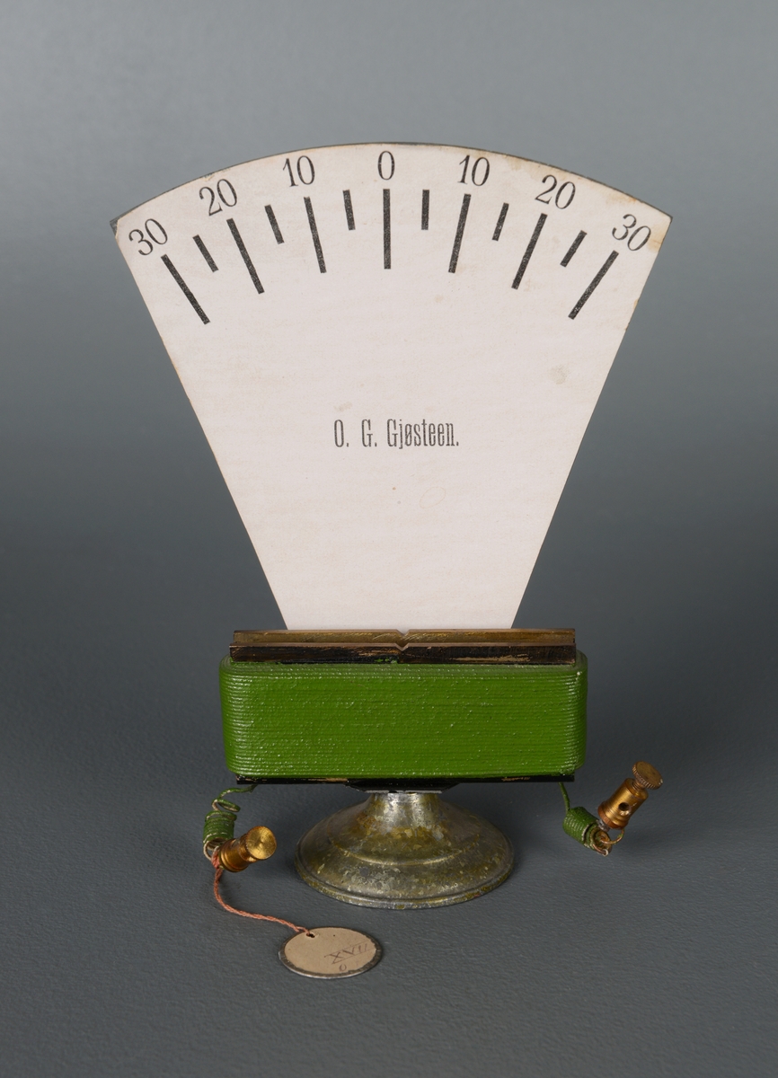 Et elektromagnetisk måleinstrument antakelig for å måle volt. En avlang spole med jernkjerne som står på en rund jernfot med hvit gradskive over. Instrumentet mangler målenålen. Gradeskalaen går fra 30 - 0 - 30. På gradskiven står det "O. G. Gjøsteen.".
