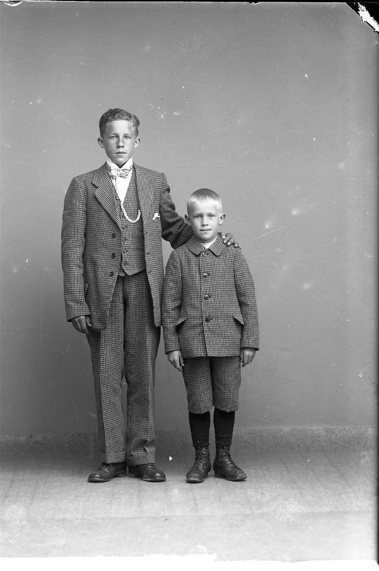 Två bröder i smårutig kostym står intill varandra. Den äldre med fluga och klockkedja håller vänster hand på lillebrors axel.