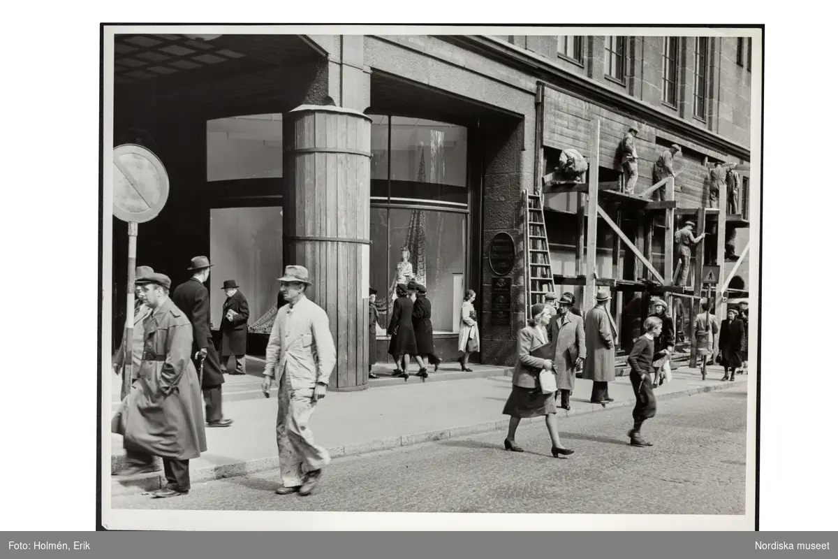 Varuhuset Nordiska Kompaniet, Hamngatan i Stockholm. På byggnadsställningar arbetar man med rivning av skyltfönstrens splitterskydd i samband med freden i maj 1945. Människor går in och ut eller passerar på trottoaren.