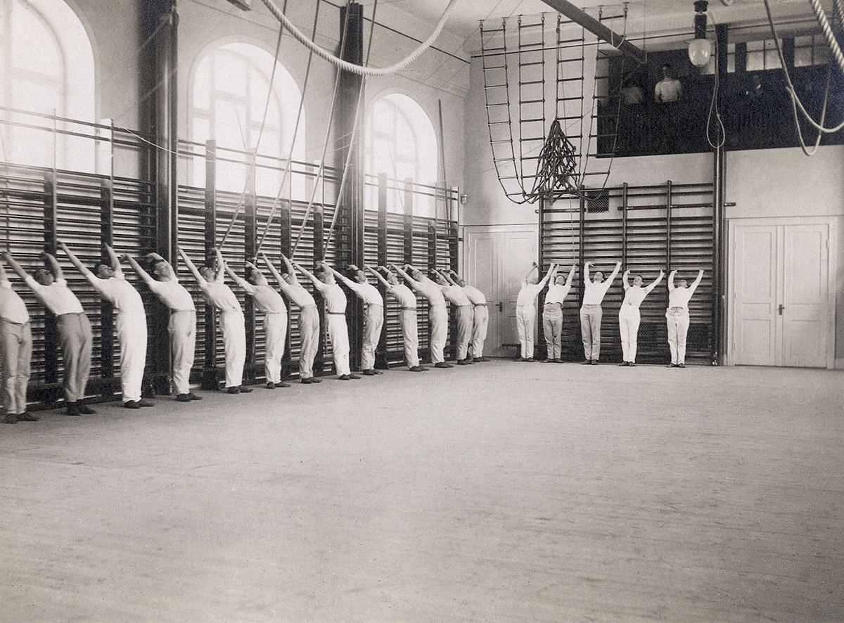 En grupp manliga gymnaster vid ribbstolarna i gymnastiksalen på Ringsbergsskolan, Växjö, 
På den lilla läktaren i fonden syns några åskådare. Ca 1914.