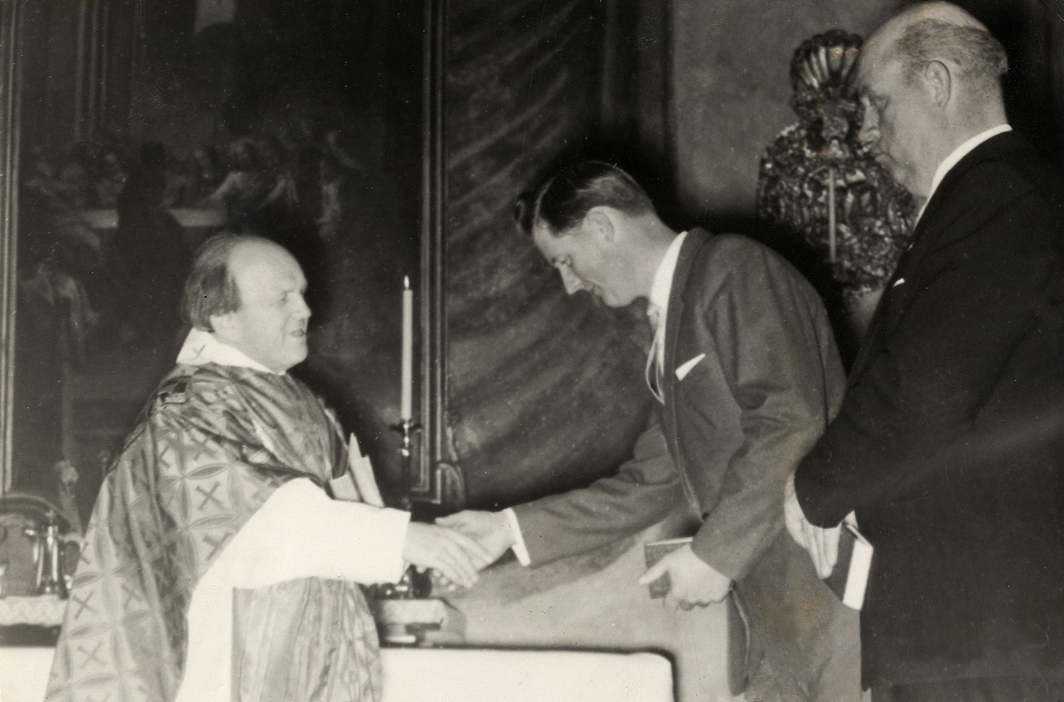 Kyrkoherde Sven Norlén, Alvesta, i fullt ornat, hälsar på en man i kostym i Aringsås kyrka. 
Ca 1965.