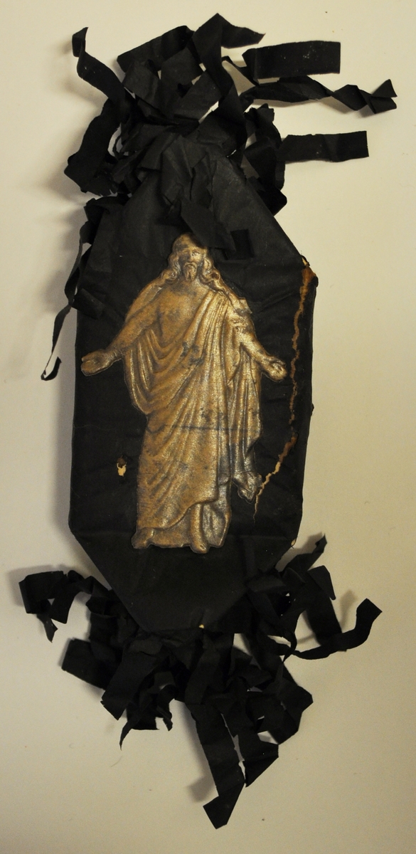 Rektangulär karamell inslagen i svart silkespapper som är krusat i ändarna. På framsidan finns ett präglat bokmärke förställande Jesus i beige färg. Skadat papper på ena sidan.