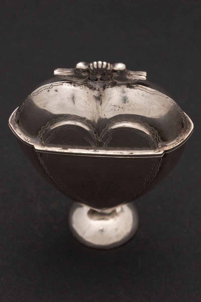 Hjerteformet luktevannshus i sølv med rund fot og to duer med kronelignede ornament mellom seg på lokket. Innvendig løs, perforert lokk. og svamp. Dekorert med prikkete bølgelinjer. Innvendig forgylt.