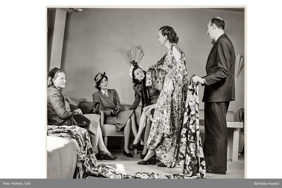 Mannekäng visar mönstrad klänning för tre kvinnliga kunder sittande i soffa. Pelle Lundgren visar ett blommigt tyg. NK:s Franska damskrädderi.