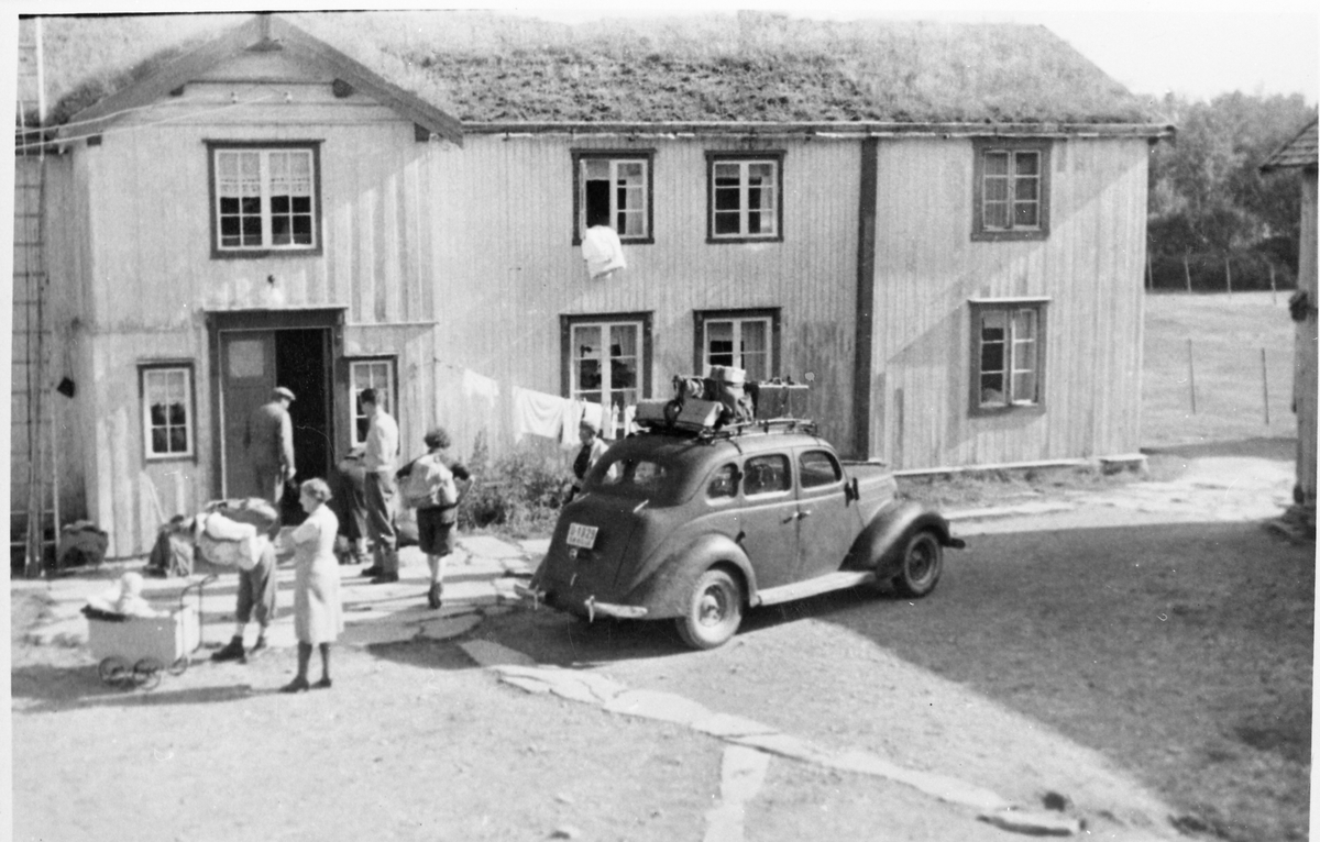 Flere folk, blant annet sommergjester, og en bil med oppakning på taket, på gårdstunet på Volldalen, sommeren 1946.