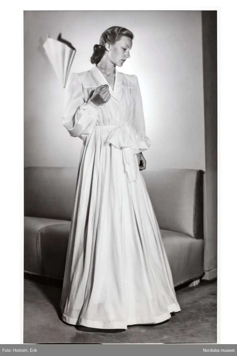 En mannekäng poserar i provrummet på NK:s Franska damskrädderi. Hon är iklädd en hellång dressinggown (morgonrock) med stor rosett i midjan. Plagget av siden är sytt av en engelsk fallskärm.
